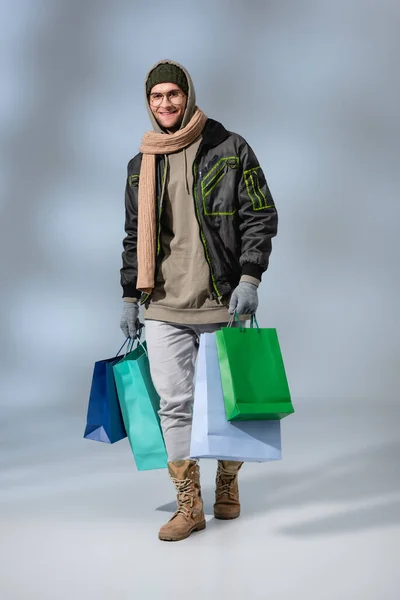 Full length of stylish man in winter outfit holding shopping bags and smiling on grey — Stock Photo
