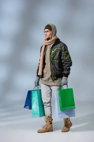Full length of young man in winter outfit holding shopping bags and walking on grey — Stock Photo