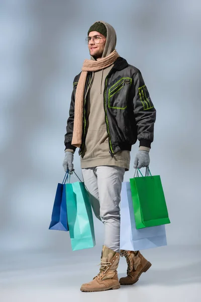 Full length of smiling man in winter outfit holding shopping bags on grey — Stock Photo