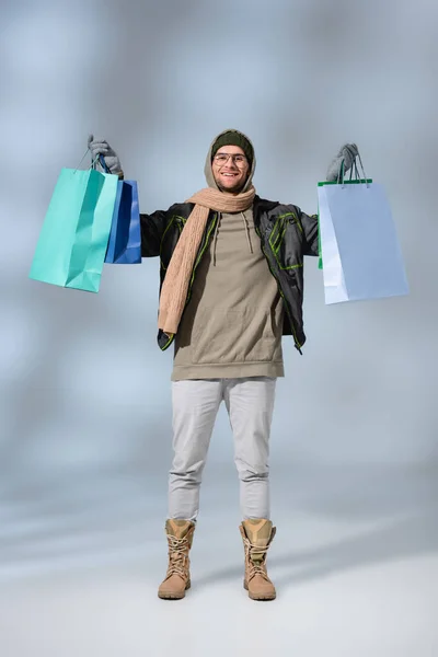 Piena lunghezza di uomo felice in parka che tiene borse della spesa su grigio — Foto stock