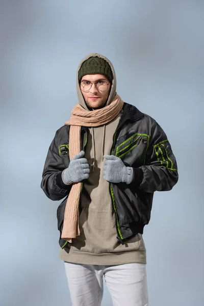 Joven hombre con estilo en sombrero, sudadera con capucha y anorak mirando a la cámara en gris - foto de stock