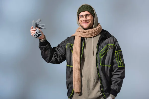 Cheerful man in parka holding gloves and smiling on grey — Stock Photo