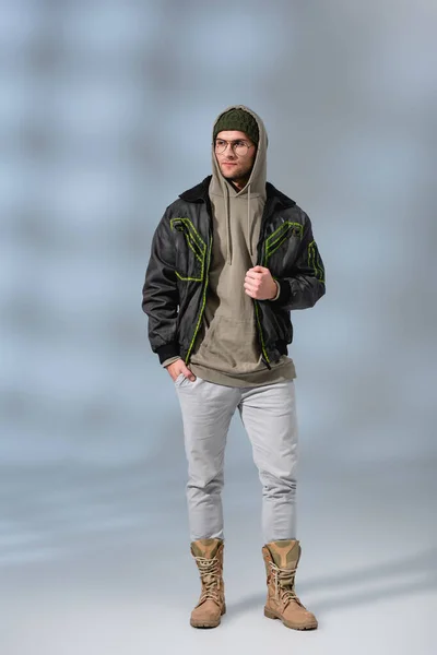 Stilvoller Mann mit Hut, Kapuzenpullover und Anorak, der mit der Hand in der Tasche auf grau steht — Stockfoto