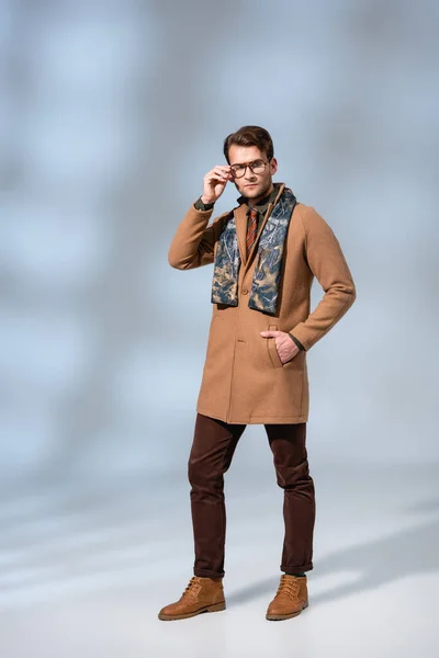 Pleine longueur de l'homme à la mode en manteau d'hiver lunettes de réglage tout en se tenant avec la main dans la poche sur le gris — Photo de stock