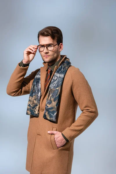 Trendy man in winter coat adjusting glasses while standing with hand in pocket on grey — Stock Photo