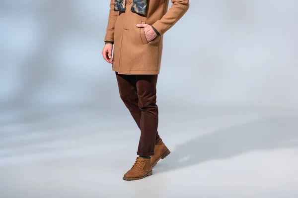 Vista recortada del hombre en abrigo de invierno y botas de pie con la mano en el bolsillo en gris - foto de stock
