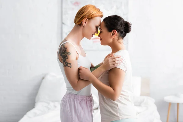 Vista lateral de mujer lesbiana tatuada abrazando hombros de novia afroamericana en el dormitorio - foto de stock