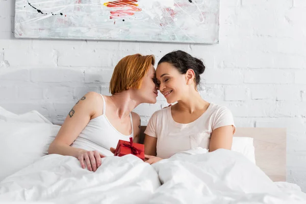 Redhead lesbian woman kissing african american girlfriend holding valentines day gift in bed — Stock Photo