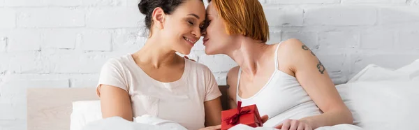 Lesbian woman kissing african american girlfriend holding gift box in bed, banner — Stock Photo