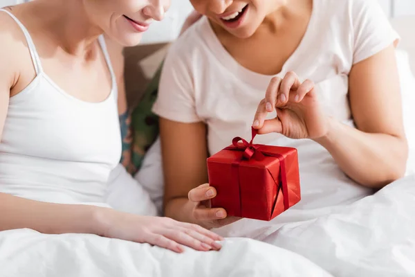 Обрізаний вид збудженої лесбіянки відкриває подарункову коробку біля дівчини в ліжку — стокове фото
