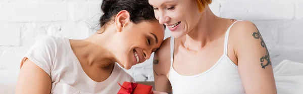 Happy lesbian interracial girlfriends smiling near gift box on valentines day in bedroom, banner — Stock Photo