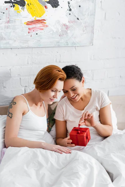 Feliz Africano americano mulher abertura caixa de presente perto ruiva lésbica namorada na cama — Fotografia de Stock