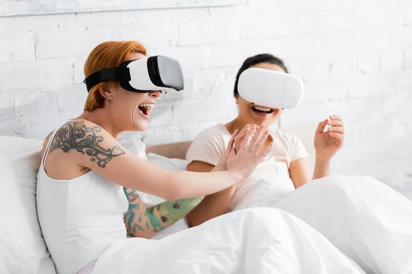Excited lesbian woman touching hand of african american girlfriend while having fun in vr headsets in bed together — Stock Photo