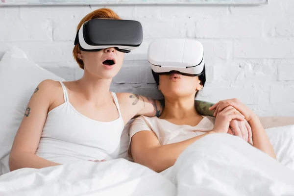 Mulher lésbica surpreso no vr headset abraçando ombro de afro americana namorada na cama — Fotografia de Stock