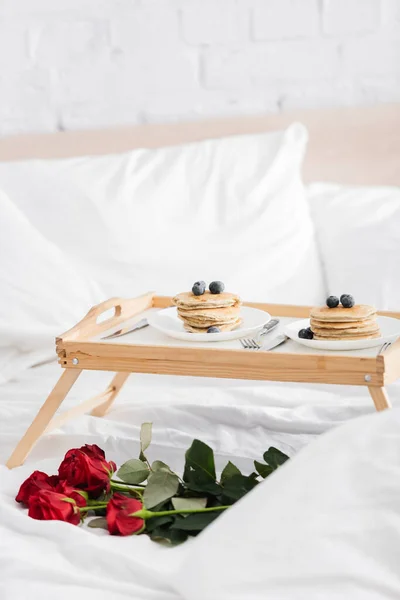 Bandeja de madera con deliciosos panqueques y arándanos cerca de rosas rojas en ropa de cama blanca, concepto de día de San Valentín - foto de stock