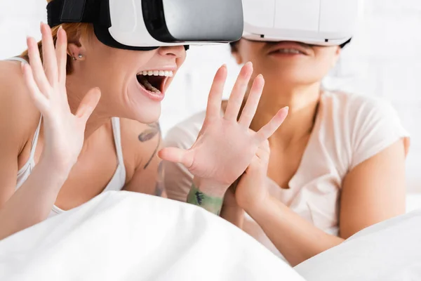 Amazed lesbian woman gesturing in vr headset near african american girlfriend in bed, blurred background — Stock Photo