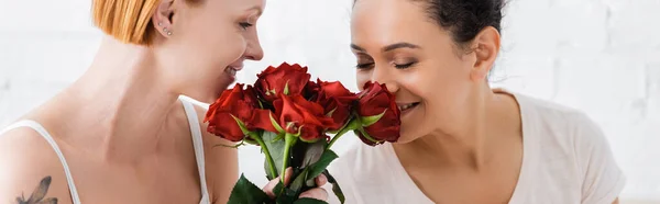 Feliz afro-americana mulher cheirando rosas vermelhas perto lésbica ruiva namorada, banner — Fotografia de Stock