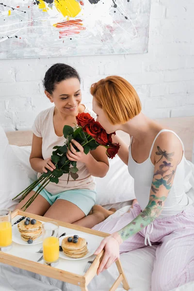 Pelirroja lesbiana mujer oliendo rosas rojas en manos de la novia afroamericana cerca de bandeja con desayuno - foto de stock