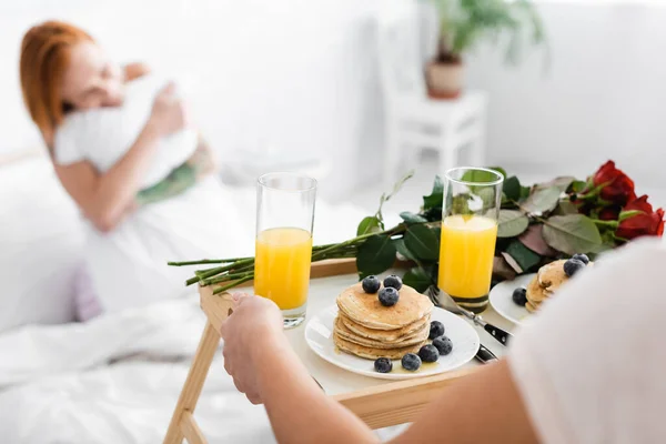 Lesbian woman holding tray with breakfast and red roses near girlfriend in bed on blurred background — Stock Photo