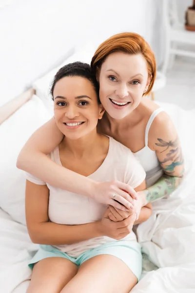 Feliz casal lésbico interracial abraçando e olhando para a câmera no quarto — Fotografia de Stock