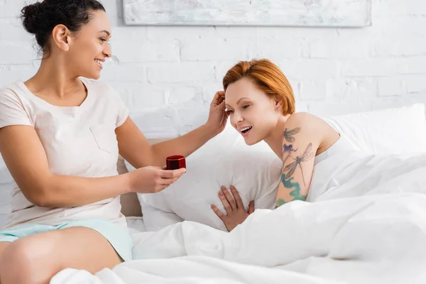 Mujer afroamericana feliz tocando el pelo de novia lesbiana asombrada mientras que hace la propuesta de boda - foto de stock