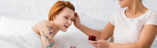 Lesbian woman touching hair of redhead girlfriend while holding jewelry box and making wedding proposal, banner — Stock Photo