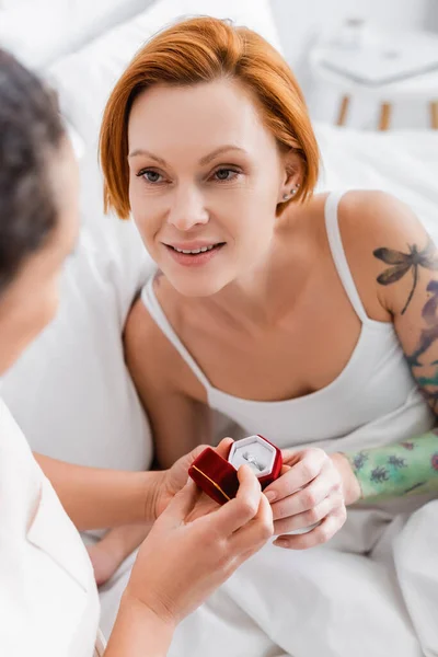 Lesbian woman holding jewelry box with ring while making wedding proposal to redhead girlfriend, blurred foreground — Stock Photo