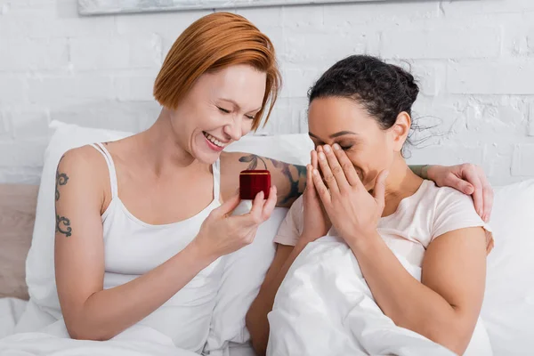 Cheerful lesbian woman holding jewelry box while making wedding proposal to amazed african american girlfriend — Stock Photo