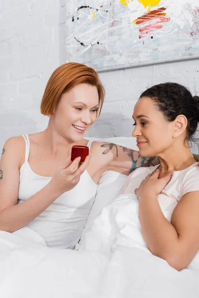 Smiling lesbian woman holding jewelry box while making wedding proposal to african american girlfriend in bed — Stock Photo