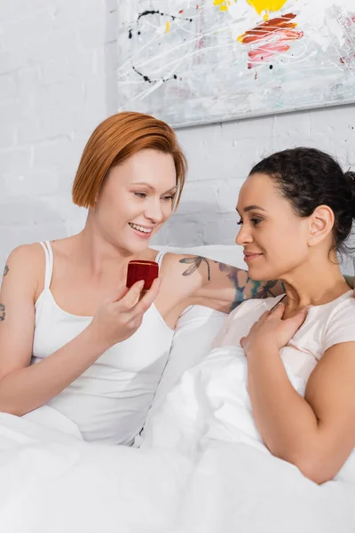 Sonriente mujer lesbiana sosteniendo joyero mientras hace propuesta de boda a la novia afroamericana en la cama - foto de stock