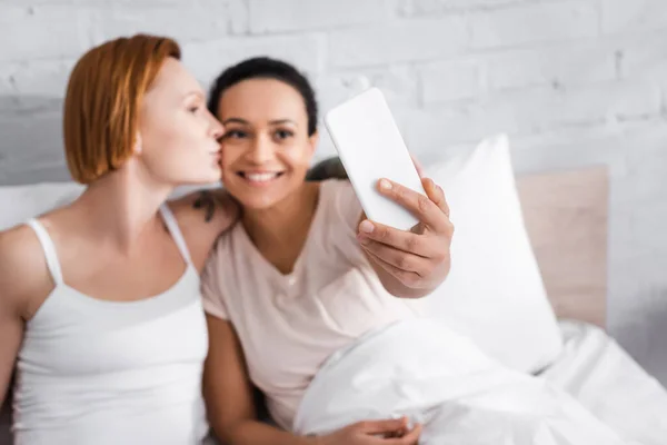 Cheerful african american woman taking selfie with redhead lesbian girlfriend kissing her in bed, blurred foreground — Stock Photo