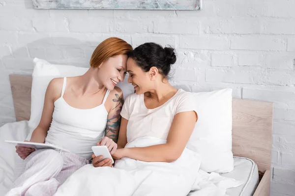 Glückliches lesbisches interracial Paar lächelt von Angesicht zu Angesicht, während es Gadgets im Bett hält — Stockfoto