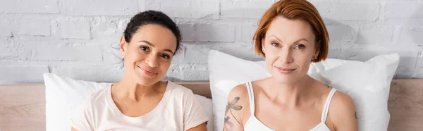 Smiling interracial lesbian couple looking at camera in bedroom, banner — Stock Photo