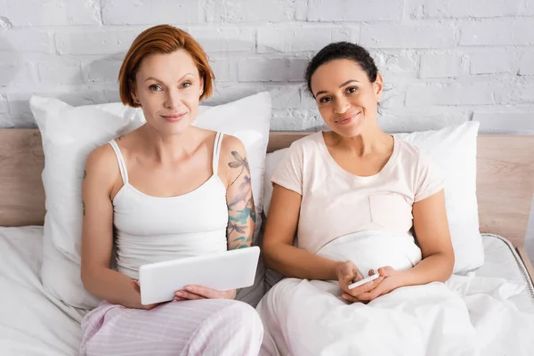 Feliz interracial lesbianas pareja mirando cámara mientras usando gadgets en la cama - foto de stock