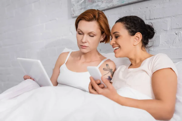 Redhead lesbian woman showing digital tablet to african american girlfriend holding smartphone in bed — Stock Photo