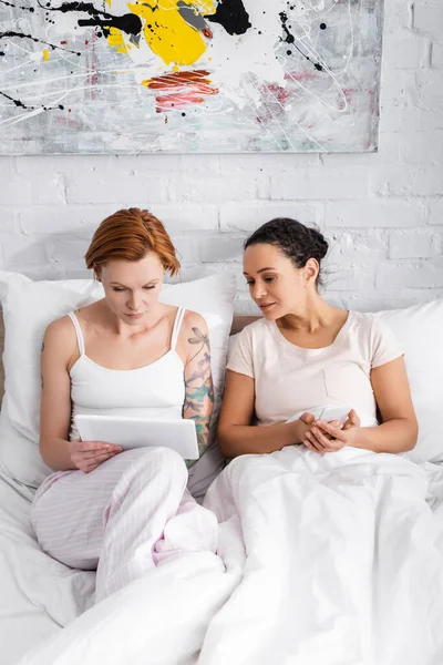 Curious african american woman holding smartphone near lesbian girlfriend using digital tablet in bed — Stock Photo