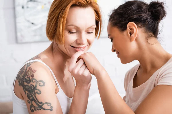 Улыбающаяся татуированная женщина трогает руку африканской девушки-лесбиянки в спальне — стоковое фото