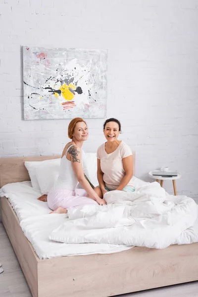 Счастливая межрасовая лесбийская пара улыбается перед камерой в спальне — стоковое фото