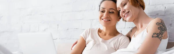 Cheerful interracial lesbian couple watching movie on computer in bed, banner — Stock Photo