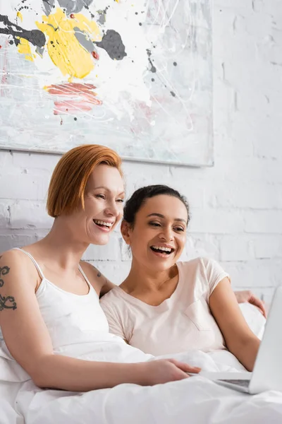 Excitado interracial lesbianas pareja riendo mientras viendo película en portátil en la cama, - foto de stock