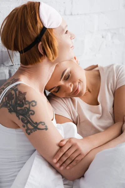 Tatoué lesbienne femme dans sommeil masque sur front embrassant afro-américaine copine au lit — Photo de stock