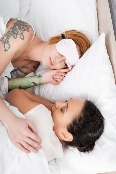 Top view of tattooed lesbian woman in eyemask hugging african american girlfriend sleeping beside in bed — Stock Photo