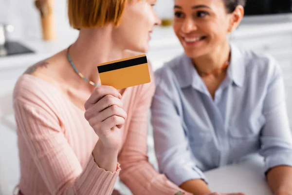 Enfoque selectivo de la tarjeta de crédito en la mano de la mujer lesbiana mirando alegre novia afroamericana, fondo borroso - foto de stock