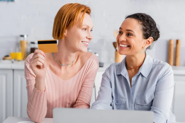 Rossa donna lesbica in possesso di carta di credito, mentre guardando sorridente ragazza africana americana in cucina — Foto stock