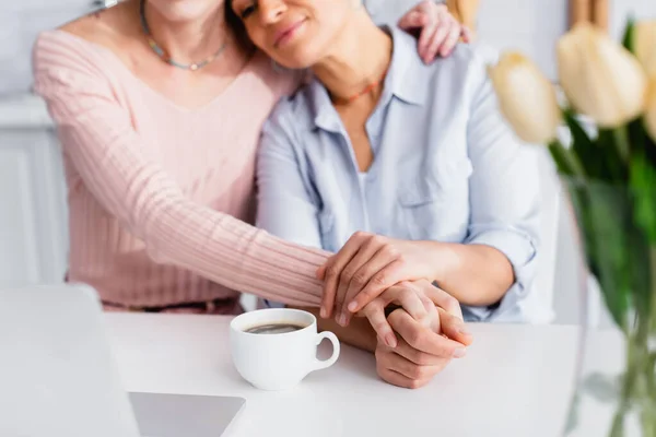 Selective focus of coffee cup near interracial lesbian couple, laptop and tulips in kitchen — Stock Photo