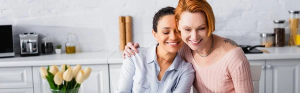 Redhead lesbian woman hugging happy african american girlfriend in kitchen near tulips on blurred foreground, banner — Stock Photo