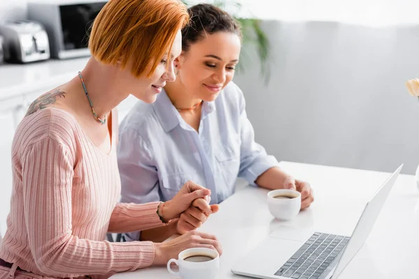 Sourire interracial lesbienne couple tenant la main près de tasses de café et ordinateur portable dans la cuisine — Photo de stock