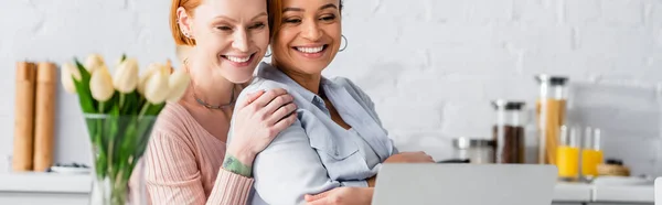 Eccitata donna lesbica che abbraccia la spalla della fidanzata afroamericana vicino a laptop e tulipani in cucina, banner — Foto stock