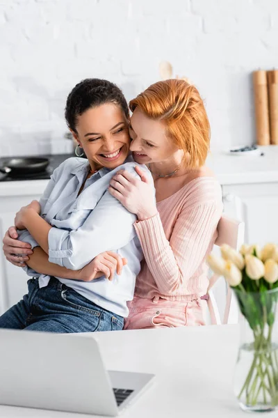 Pelirroja lesbiana mujer abrazando alegre africana americana novia cerca de portátil y tulipanes en borrosa primer plano - foto de stock