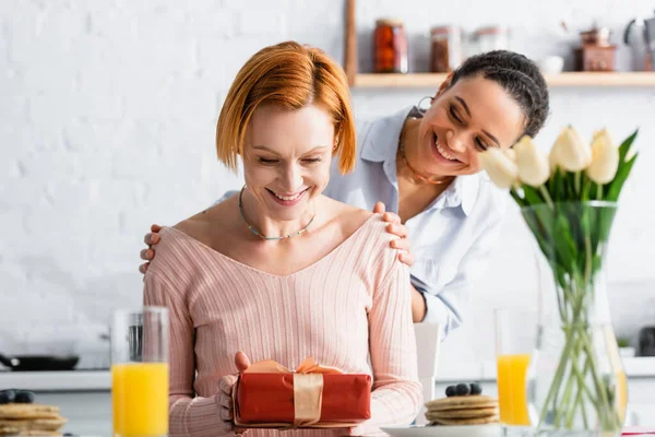 Smiling african woman hugging shoulders of happy lesbian girlfriend holding gift on valentines day, blurred foreground — Stock Photo